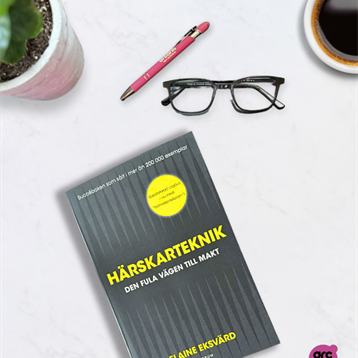 Bild på en bok på ett bord med glasögon, kaffekopp, penna och en blomma